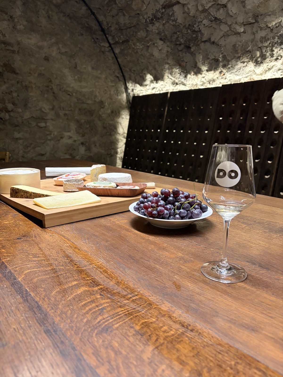 Verre gravé DO, planche de fromages et raisin sur la grande table en chêne de la cave voûtée du domaine